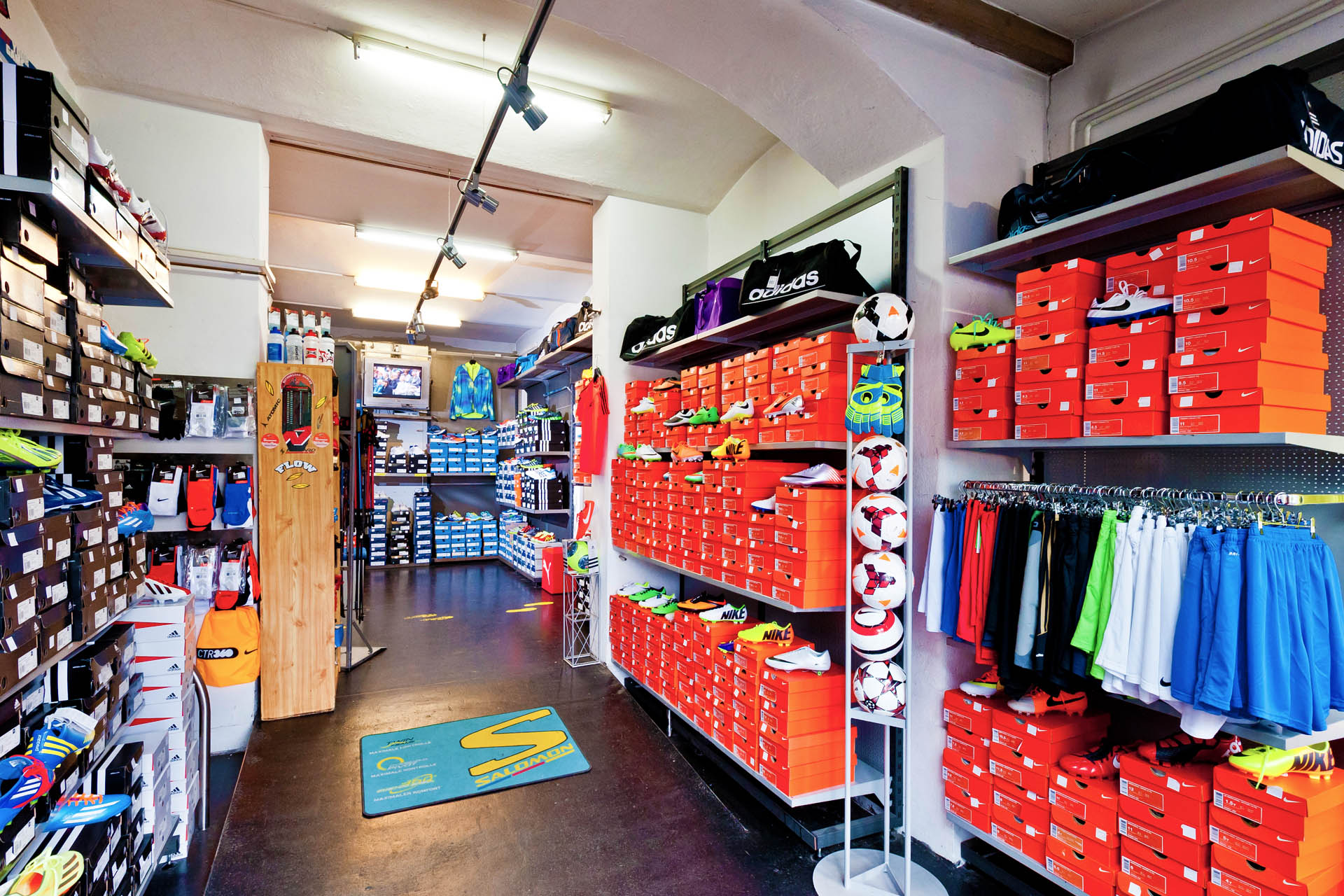 Sportgeschäft mit Regalen voller Sportartikel, darunter Schuhe und Bekleidung in orangefarbenen Boxen und verschiedenen Farben.