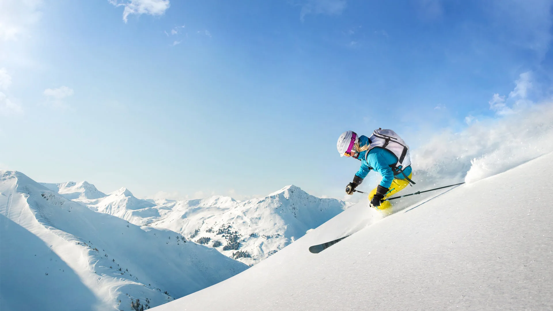 Skifahrer in blauer Jacke und gelben Hosen fährt eine verschneite Piste in den Alpen hinunter, umgeben von schneebedeckten Bergen.